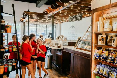 A group of young women in red shirts chat near a coffee shop counter, surrounded by snacks and merchandise displays.