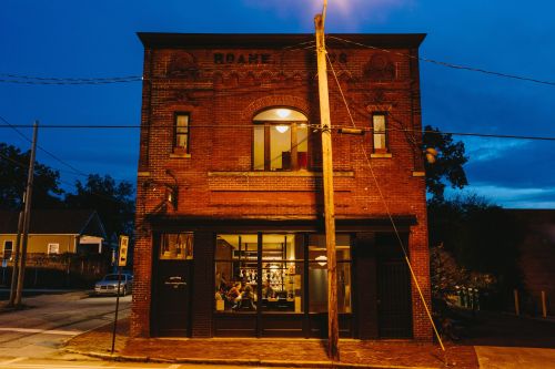 A brick building at dusk, featuring large windows and a warm glow inside, with streetlights illuminating the scene.