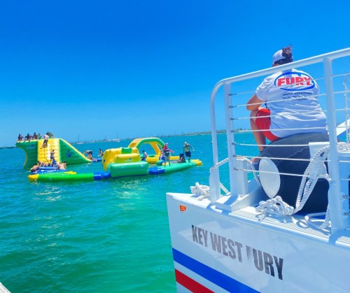 A person sits on a boat, watching people play on inflatable water attractions in clear blue water under a sunny sky.