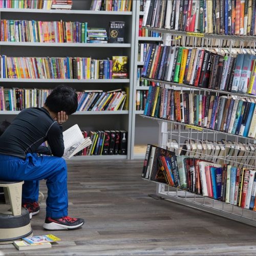A child sits on a stool, reading a book in a library filled with colorful bookshelves.