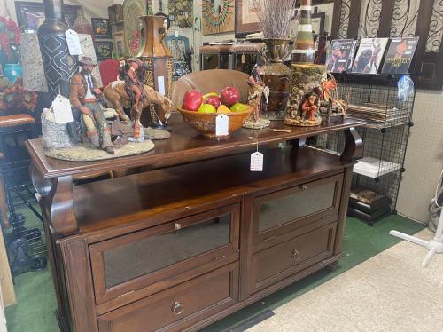 A wooden display table with decorative figurines, a bowl of apples, and magazines in a shop setting.