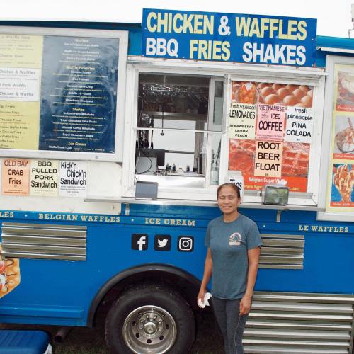 A woman stands in front of a colorful food truck offering chicken, waffles, BBQ, fries, and shakes.