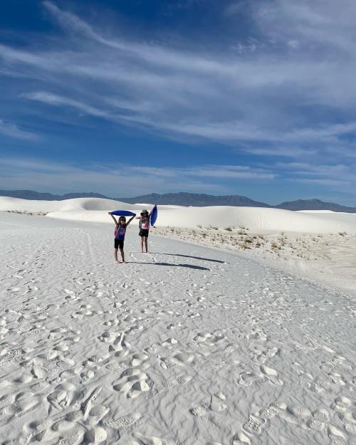 Two children play on a sandy white dune, holding blue sleds under a clear blue sky with distant mountains.