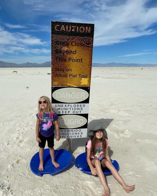 Two girls sit on blue discs in a desert area near a caution sign about unexploded munitions. Bright sky in the background.