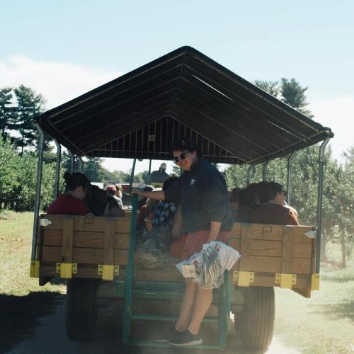 A group of people sitting on a hayride wagon in an orchard, with one person smiling and holding papers.