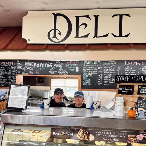 Two smiling deli workers stand behind the counter, with a menu board and deli sign above them.
