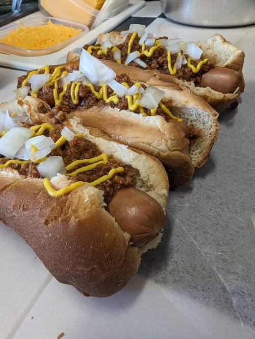 Three hot dogs in buns topped with chili, mustard, and diced onions, arranged on a countertop.