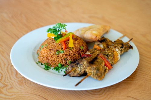 A plate of seasoned rice with vegetables, skewers of grilled meat, and a pastry, garnished with herbs.