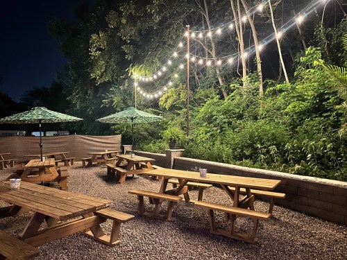 Outdoor seating area at night, featuring wooden picnic tables, string lights, and green umbrellas surrounded by greenery.