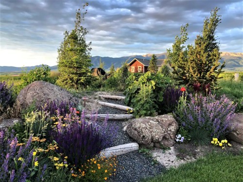 A scenic garden with colorful flowers, stone pathways, and a house in the background against a cloudy sky and mountains.