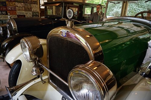 Close-up of a vintage green and white car with shiny chrome details, set in a garage with other classic cars in the background.