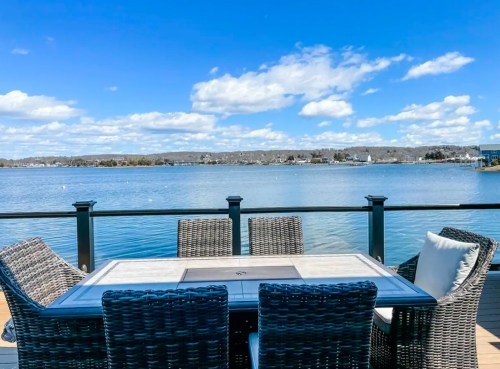 A waterfront view featuring a table and chairs on a deck, with blue skies and clouds reflected in the water.