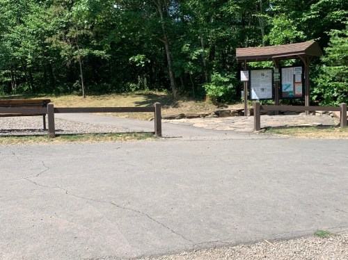 Trailhead entrance with a signboard, surrounded by trees and gravel paths. Benches are visible nearby.
