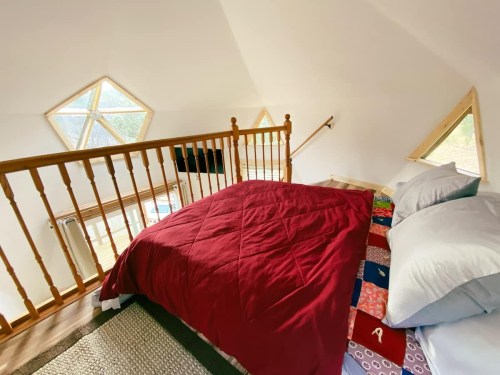 Cozy loft bedroom with a red quilt, wooden railing, and unique triangular windows letting in natural light.