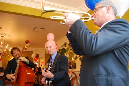 A jazz band performs in a festive setting, featuring a trumpet player, guitarist, and bassist with balloons in the background.