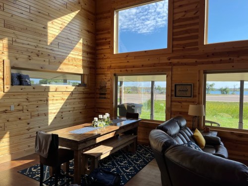 Bright, wooden interior of a cozy living space with a dining table, sofa, and large windows showcasing a scenic view.