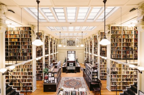 A spacious library with tall bookshelves, large windows, and a central reading area, filled with natural light.