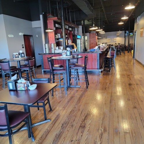 Interior of a restaurant featuring wooden floors, tables, and a bar area with a staff member in the background.