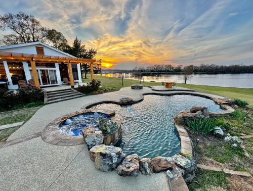 A scenic backyard with a pool, stone features, and a sunset over a lake, framed by a cozy house and greenery.
