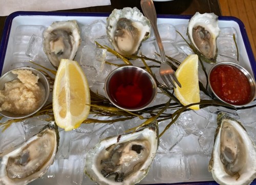 A platter of fresh oysters on ice, garnished with lemon wedges, cocktail sauce, and horseradish.