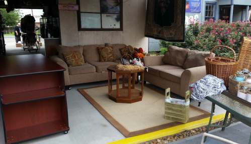 A cozy outdoor seating area with a sofa, coffee table, and decorative items, surrounded by greenery and a rustic setting.