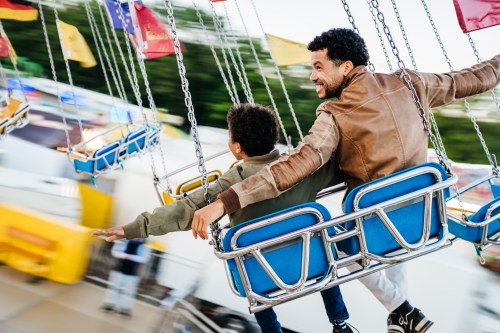 A single dad sitting on a spinning swing ride with his son, both of them with their arms stretched out while spending the day at the fun fair.