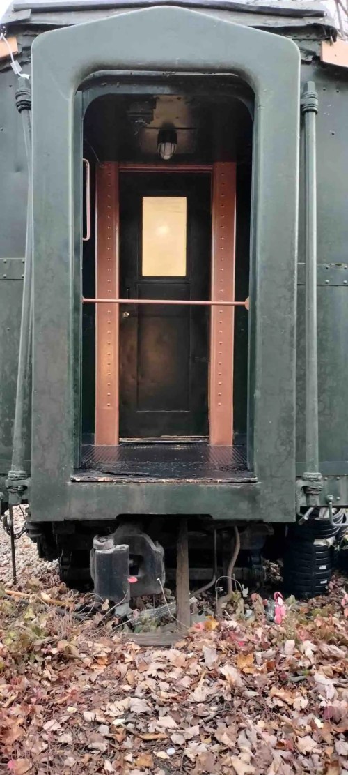A green train car with an open door, revealing a small interior space and a window, surrounded by fallen leaves.