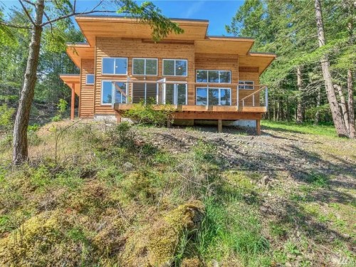 Modern wooden house with large windows, set on a hillside surrounded by trees and greenery.