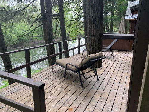 A wooden deck by a river, featuring a lounge chair and a hot tub, surrounded by trees.