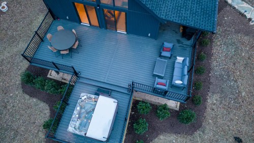Aerial view of a modern deck with a hot tub, seating area, and dining table surrounded by greenery.