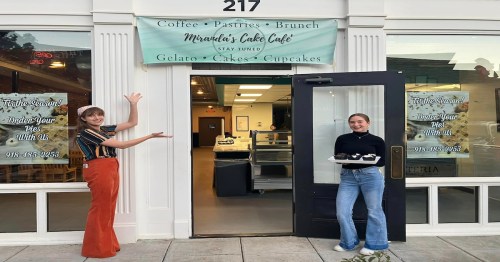 Two women stand outside Miranda's Cake Café, showcasing cupcakes and welcoming customers to the café.