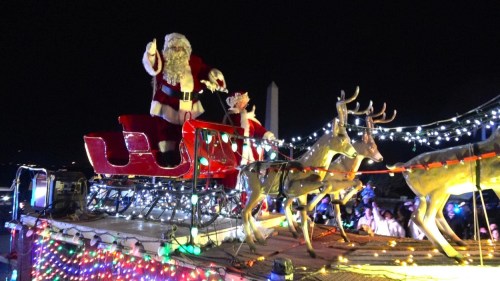 Santa Claus waves from a decorated sleigh pulled by reindeer, surrounded by colorful lights at night.