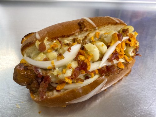 A hot dog topped with macaroni, bacon bits, onions, and crunchy toppings, served in a toasted bun.