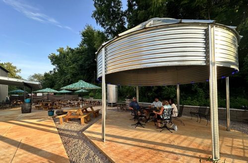 Outdoor seating area with metal structure, picnic tables, and umbrellas, surrounded by greenery and a clear blue sky.