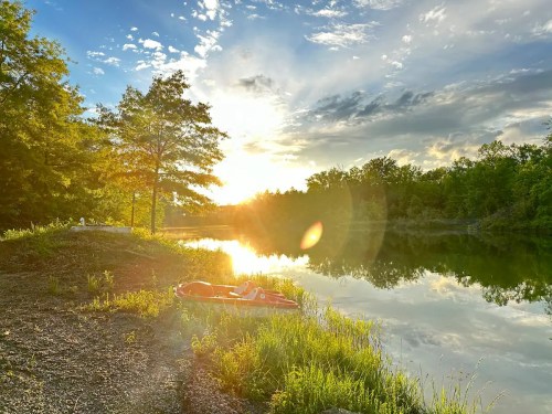 A serene lakeside scene at sunset, with a kayak on the shore and trees reflecting in the calm water.