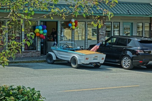 A vintage car with a wooden back and an American flag parked outside a building decorated with colorful balloons.