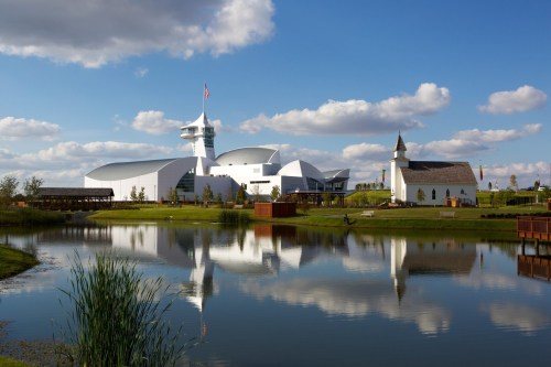 Modern buildings and a church reflect in a calm pond, surrounded by green grass and blue skies with fluffy clouds.