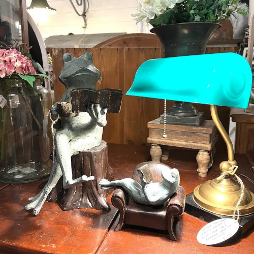 A whimsical frog sculpture reading on a log, with another frog lounging on a chair beside a vintage lamp.