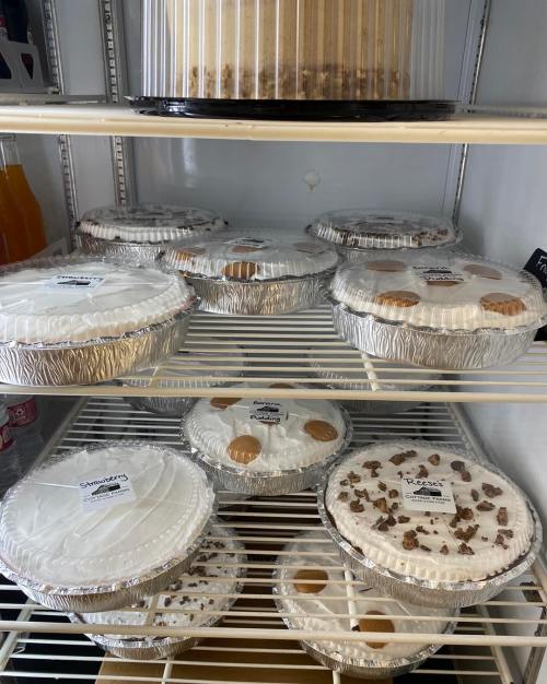 A refrigerator shelf filled with various pies in aluminum pans, labeled with different flavors.