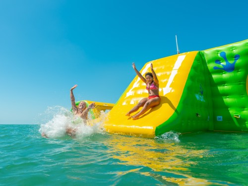 Two people enjoy a water slide on an inflatable structure in the ocean, splashing and having fun under a clear blue sky.