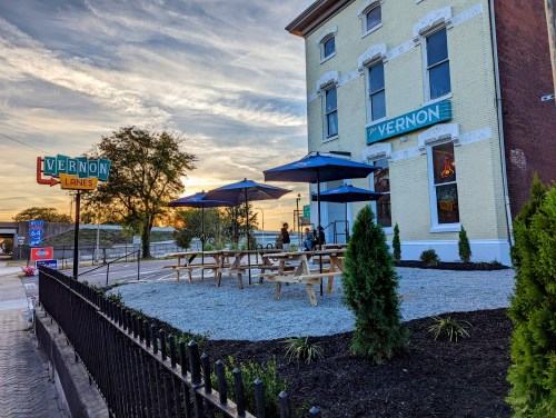 Exterior of Vernon Lanes with outdoor seating, umbrellas, and a sunset in the background. Greenery and a fence visible.
