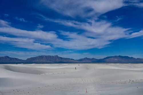 A vast white sand dune landscape under a blue sky with scattered clouds and distant mountains.