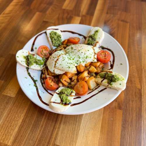 A plate of burrata cheese with cherry tomatoes, roasted potatoes, and drizzles of pesto and balsamic glaze on a wooden table.