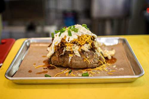 A loaded baked potato topped with cheese, sour cream, green onions, and pulled pork on a metal tray.