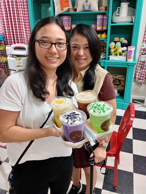 Two women hold four colorful drinks in a cozy café setting with a vibrant background.