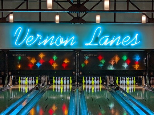 Neon sign reading "Vernon Lanes" above bowling lanes with colorful lights and pins arranged in a bowling alley.