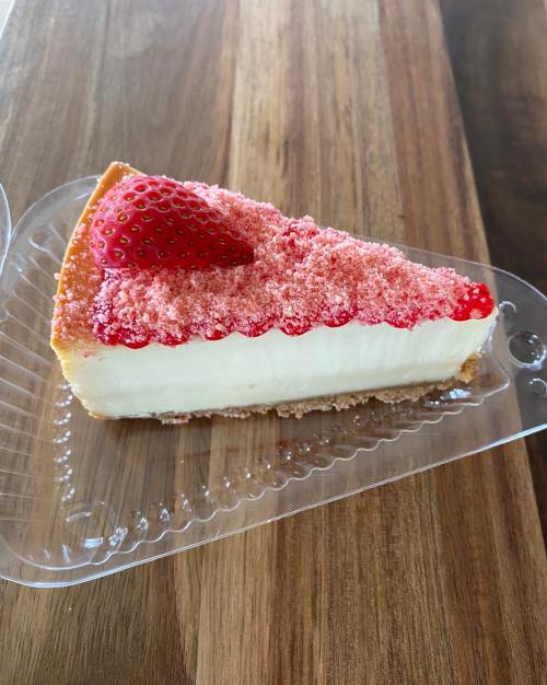 A slice of cheesecake topped with crushed strawberries and a whole strawberry, served in a clear plastic container.