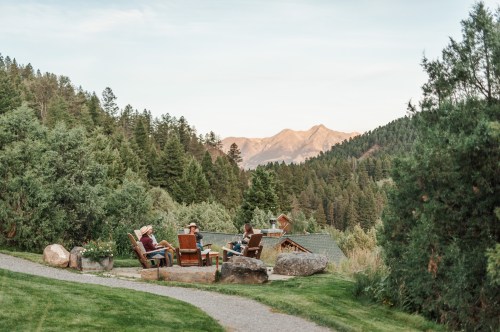 A group of people relaxes in chairs outdoors, surrounded by trees and mountains under a soft sunset sky.