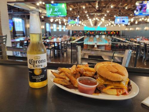 A burger with cheese and fries beside a Corona beer, set in a lively restaurant with a bowling alley in the background.