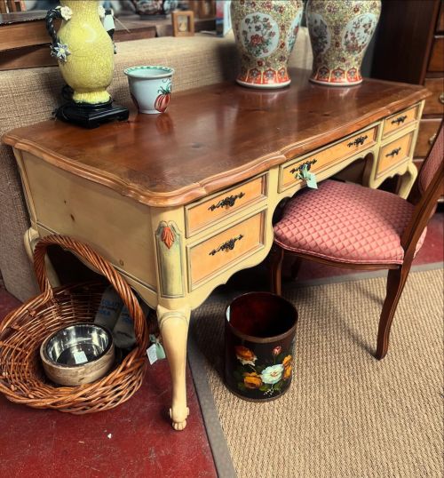 A vintage wooden desk with a chair, decorative vases, a basket, and a small trash can in a cozy interior setting.